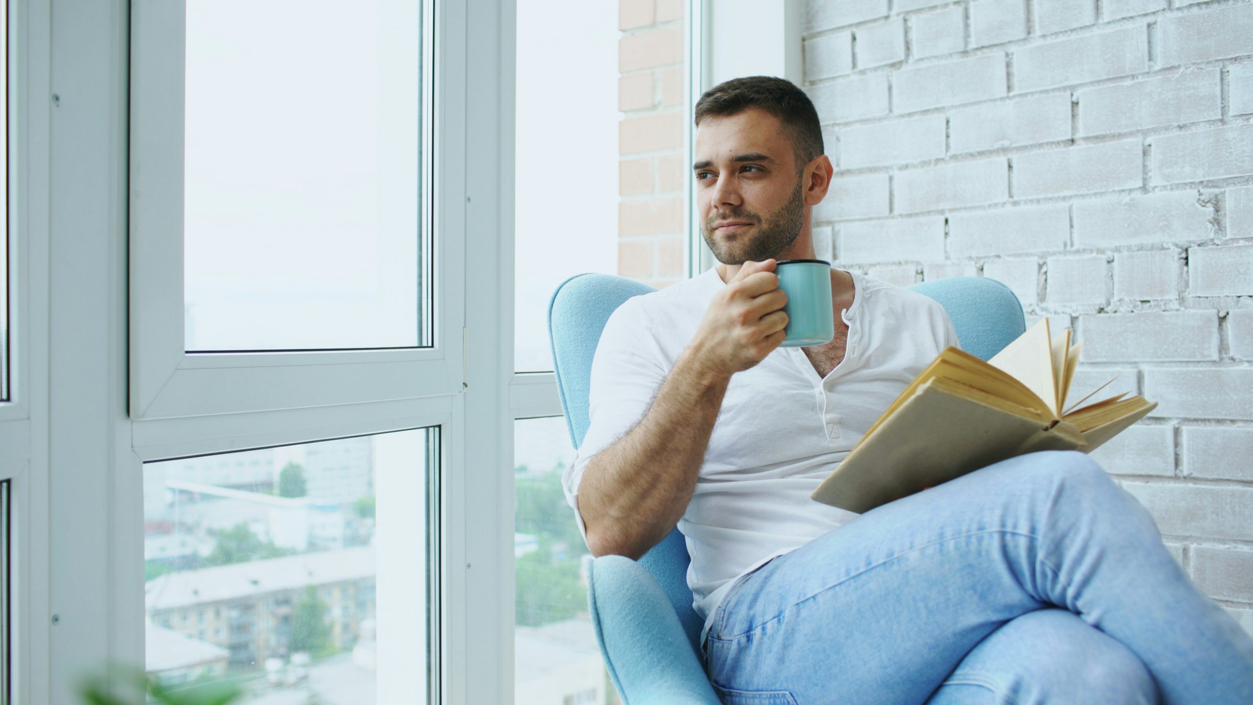 smiling man looking out a window, holding a cup and a book